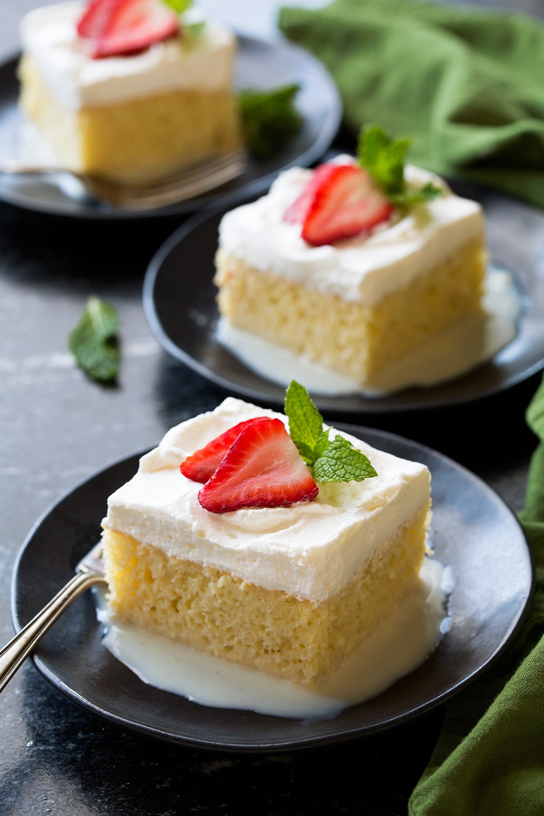 Three slices of tres leches cake on dark dessert plates set over a dark granite surface.