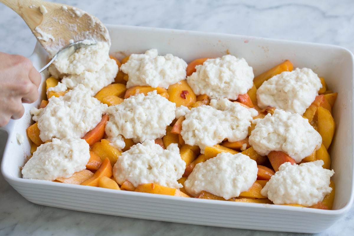 dolloping cobbler topping over peach layer in baking dish.