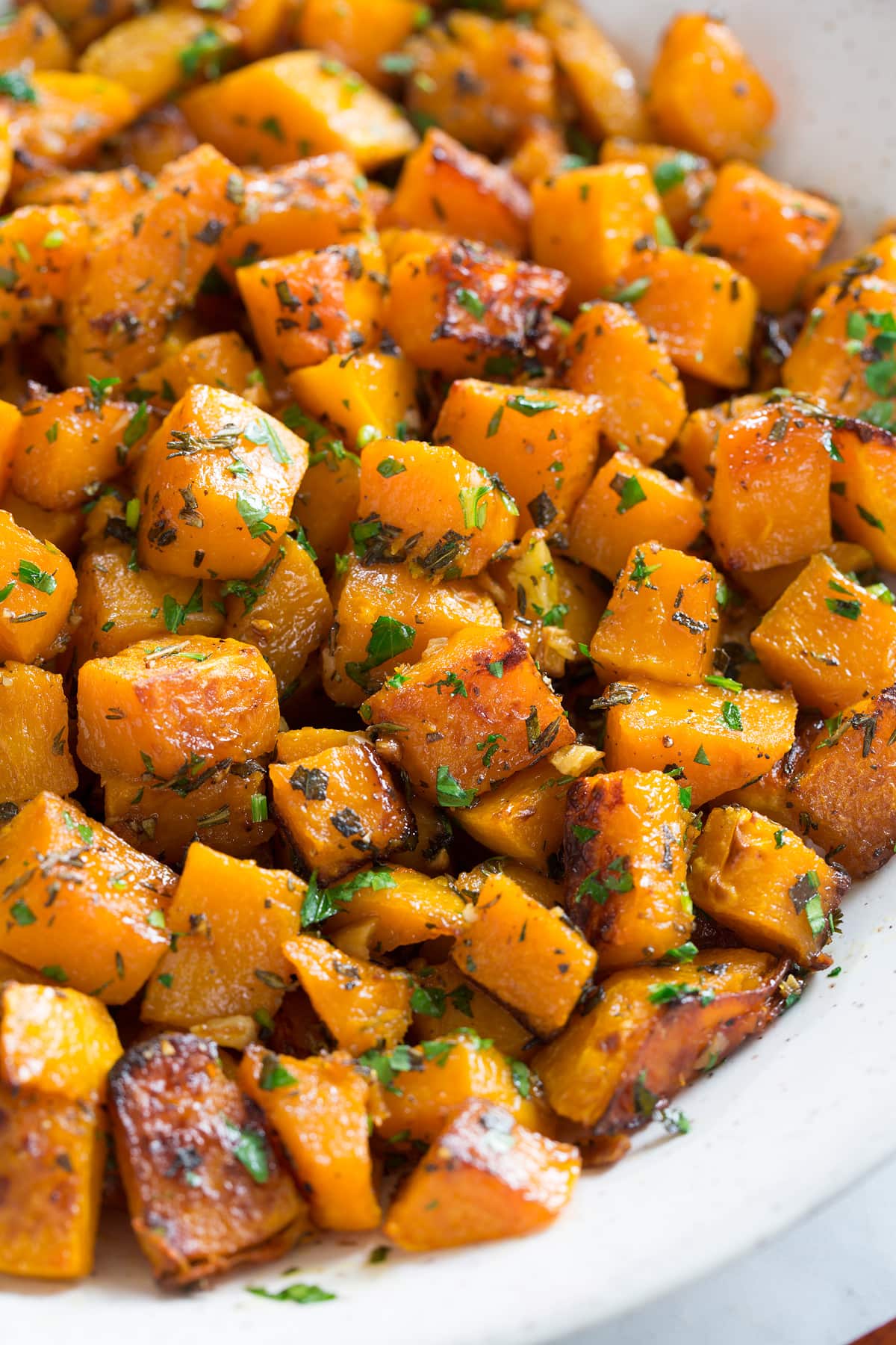 Roasted butternut squash with garlic and fresh herbs in a white ceramic serving bowl set over a white marble surface.