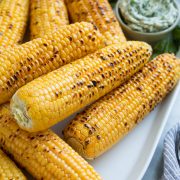 Close up image of stack of grilled corn on the cob on a white oval serving platter set over a marble surface.