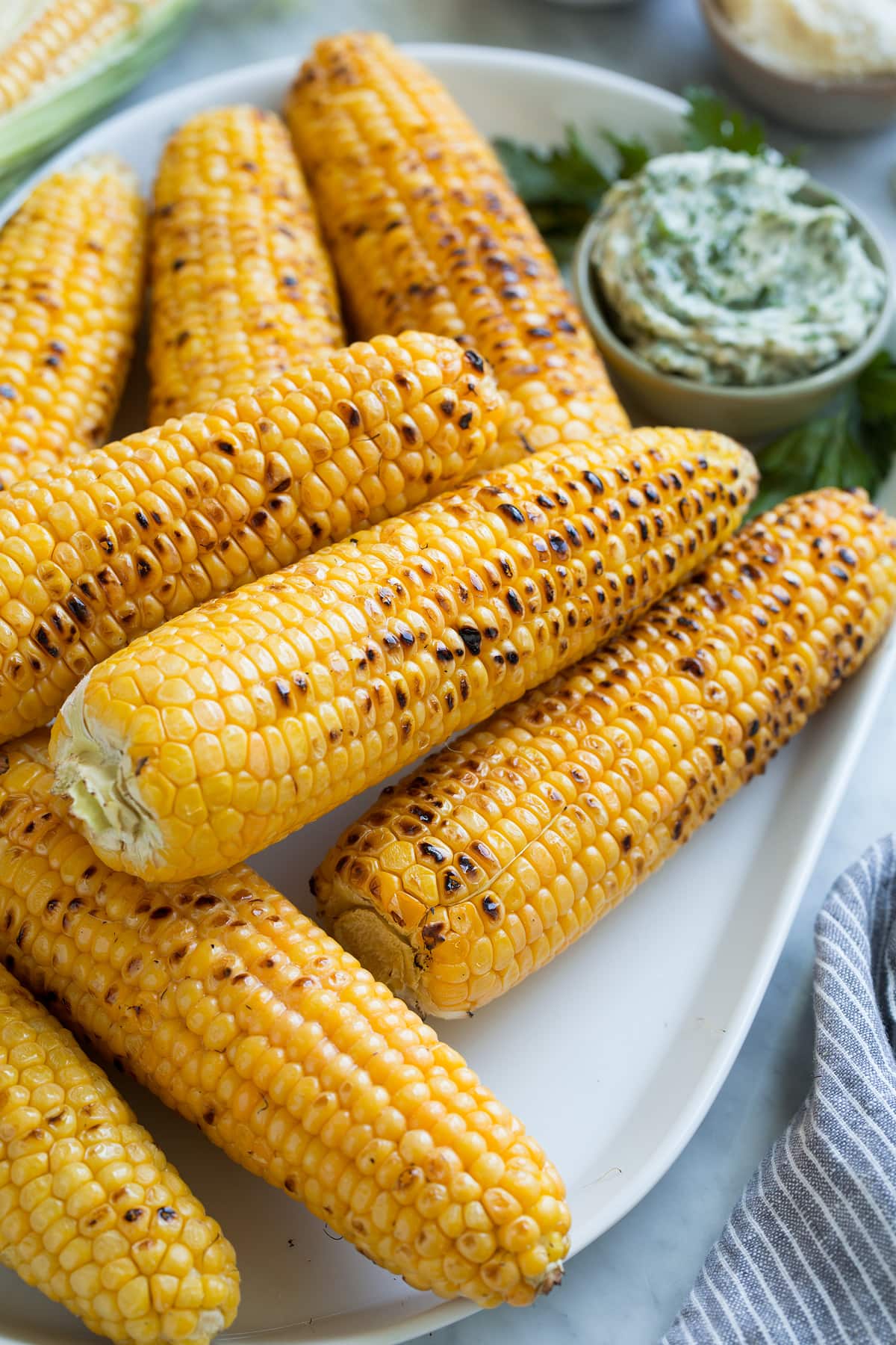 Grilled Corn on the Cob + 3 Flavored Butters Close up image of stack of grilled corn on the cob on a white oval serving platter set over a marble surface.