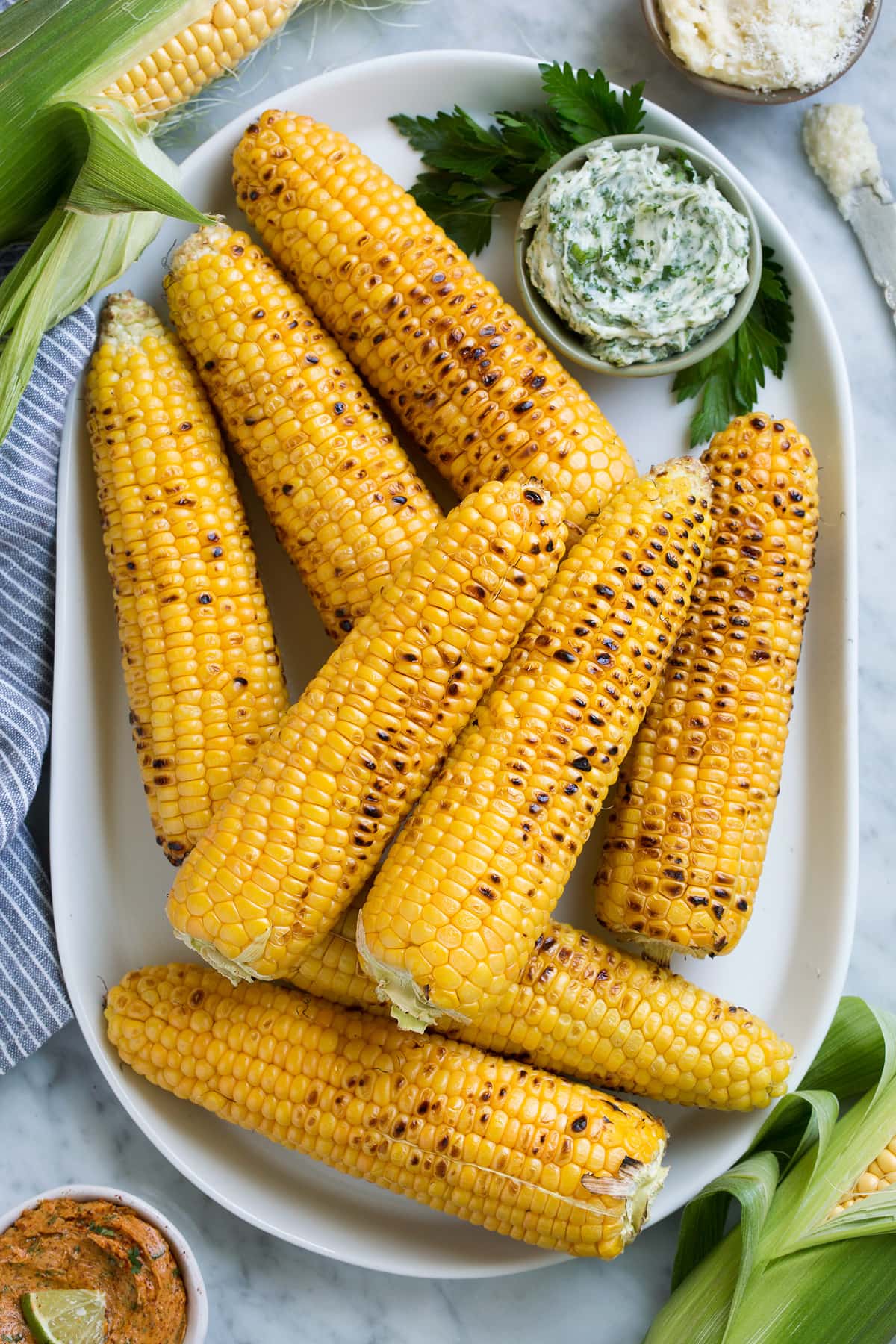 Grilled Corn on the Cob + 3 Flavored Butters Eight grilled corn on the cob on a white oval platter with flavored butters on the side.