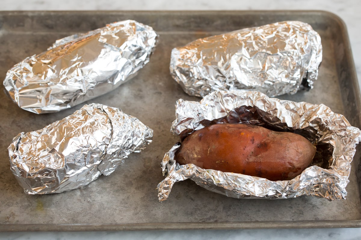 Mashed Sweet Potatoes - one of the best fall side dishes! These really are the best mashed sweet potatoes the secret ingredient is the browned butter! #mashedpotatoes #sweetpotatoes #sidedish #thanksigving Four sweet potatoes wrapped in aluminum foil, one open, sitting on a baking sheet.
