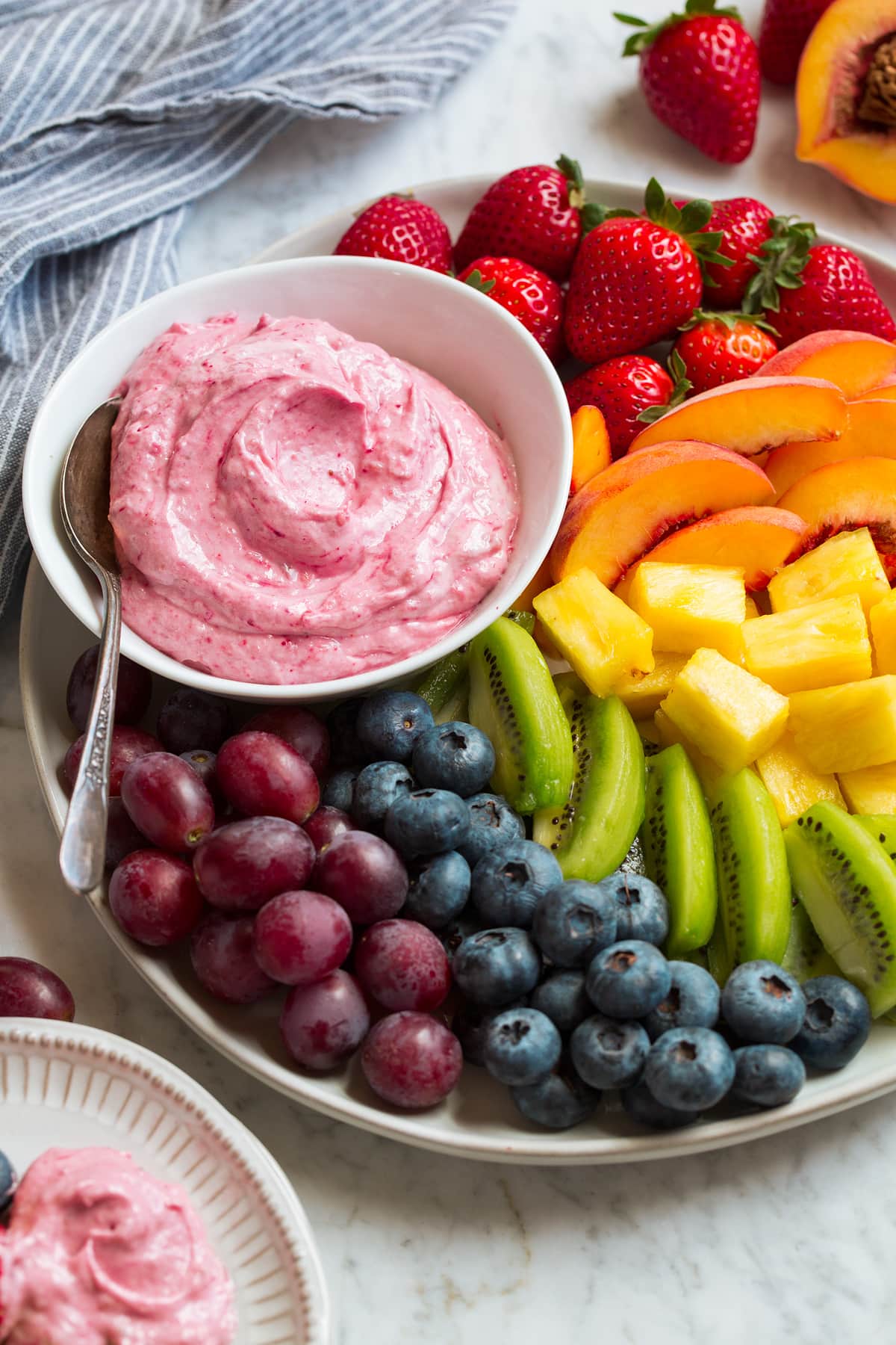 Cream cheese fruit dip in a white bowl served on a platter with fresh strawberries, peaches, pineapple, kiwi, blueberries and grapes.
