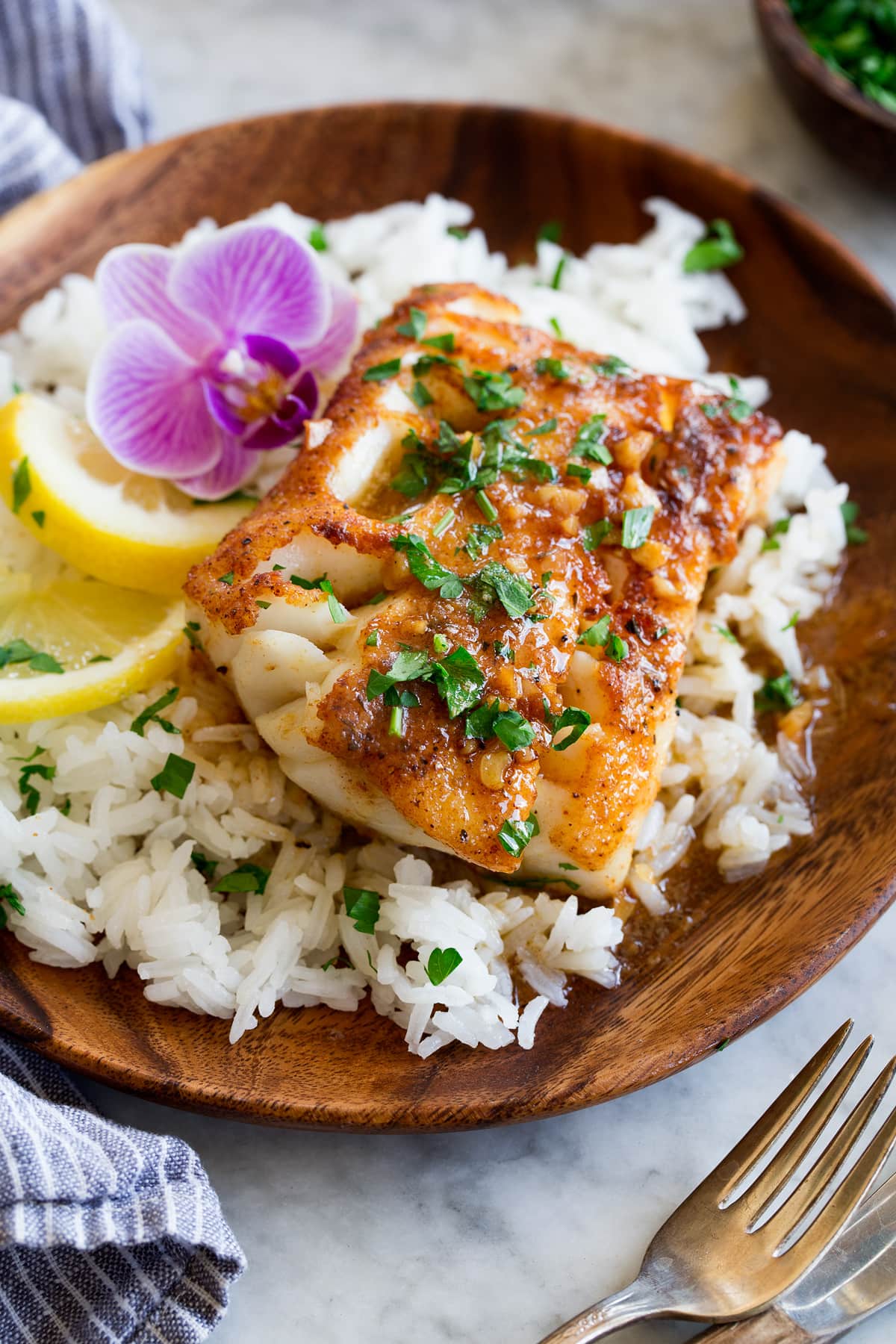 Photo: Pan seared cod fillet topped with a spice rub, lemon butter sauce and parsley. It is served over white rice with a purple flour and lemon slices to the side.