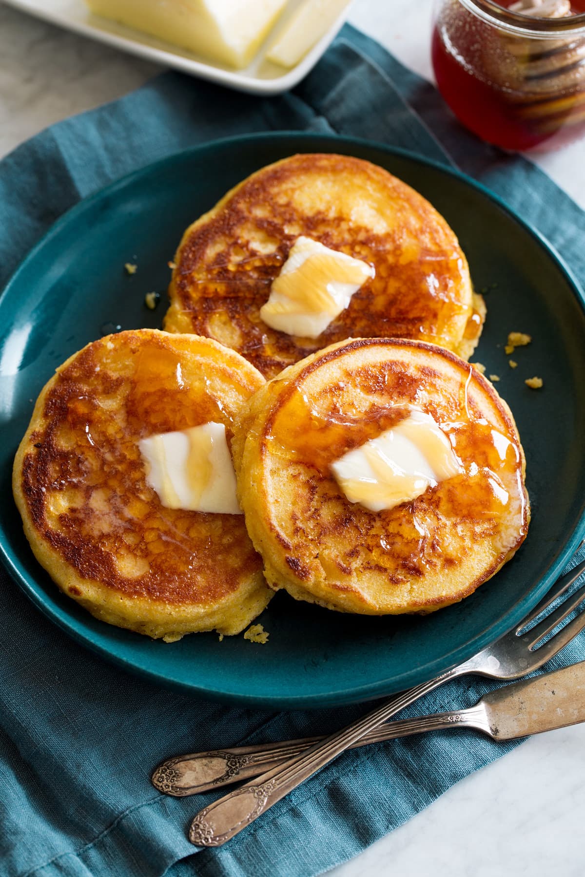 Johnny Cakes Three johnny cakes covered with dabs of butter and honey. They are a blue plate set over a blue cloth with silverware to the side.