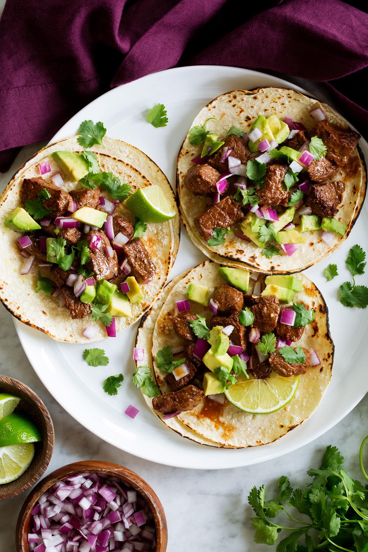 Steak Tacos Three steak tacos on a white serving plate with a purple cloth to the side.