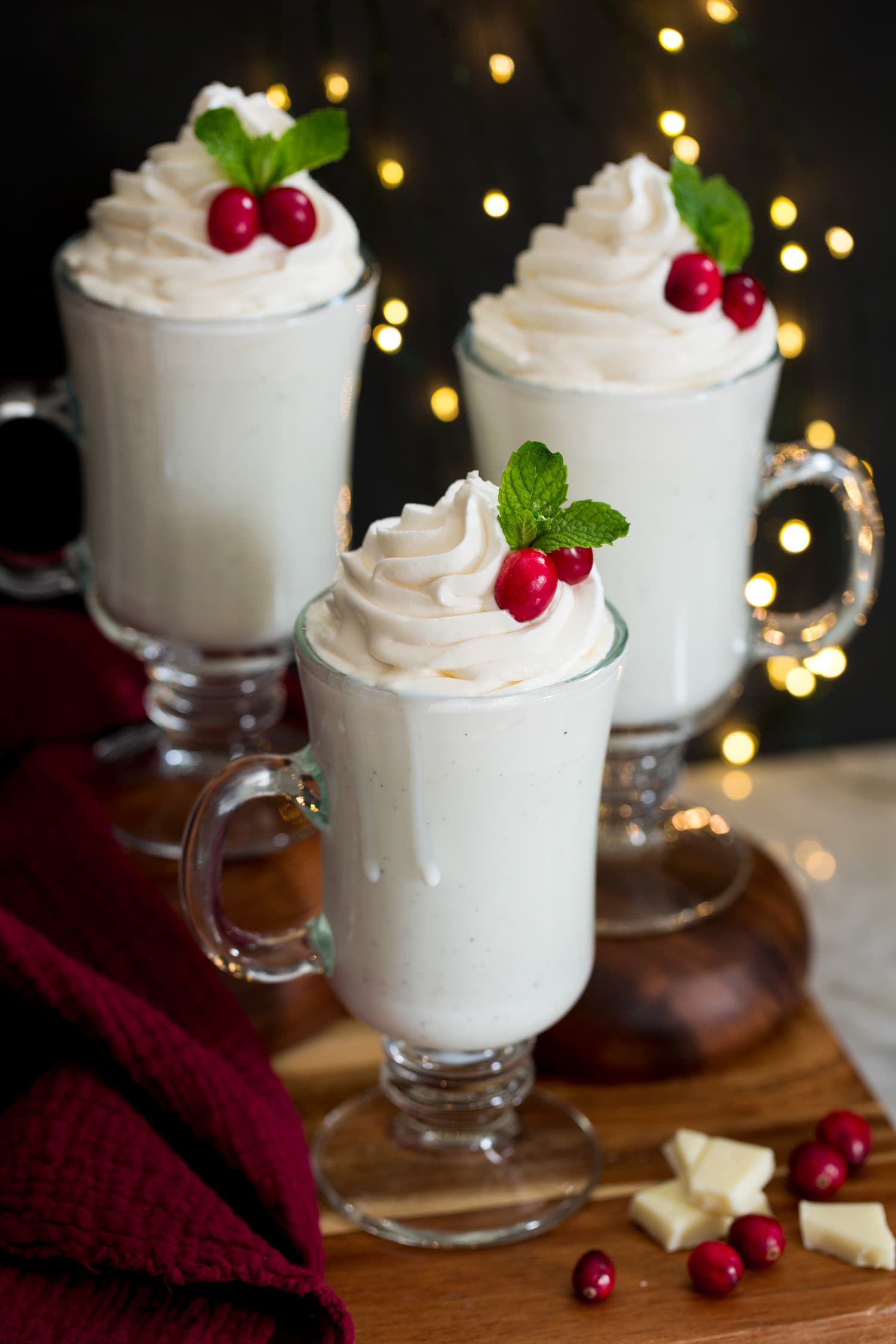White Hot Chocolate Three mugs of white hot chocolate on a wooden tray with Christmas lights in the background.