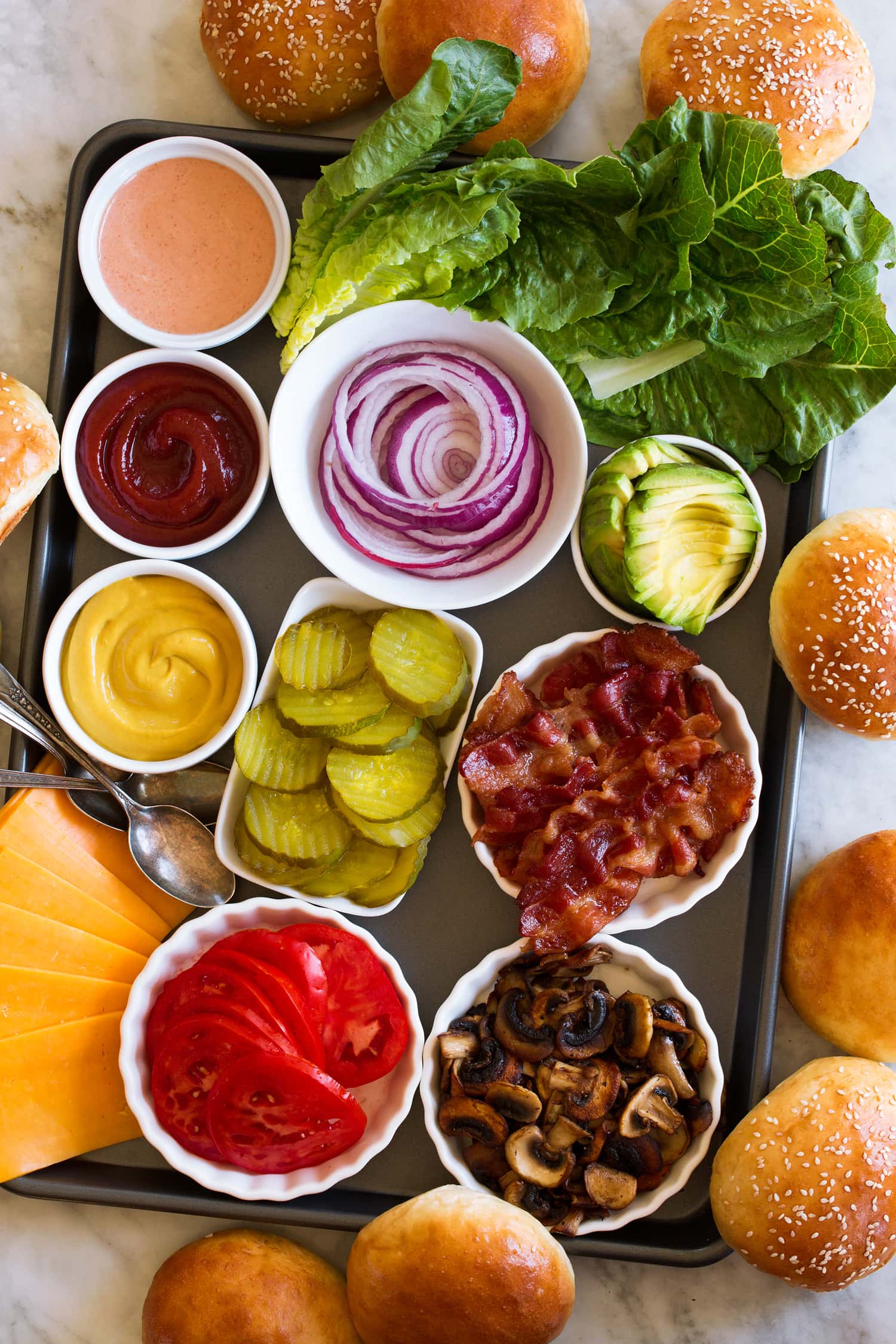 Hamburger Toppings Topping ideas for hamburgers shown on a baking sheet with hamburger buns surrounding them.