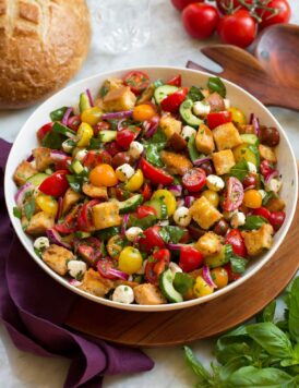 Panzanella salad in a white bowl over a wooden tray with a purple cloth and fresh basil and tomatoes shown to the sides.
