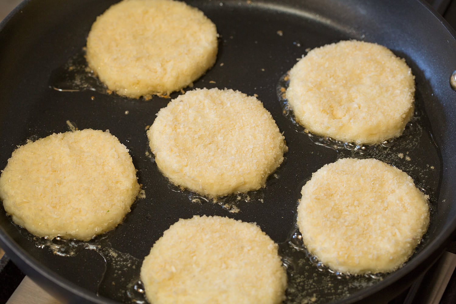Potato cakes being fried in oil in a dark non-stick skillet.