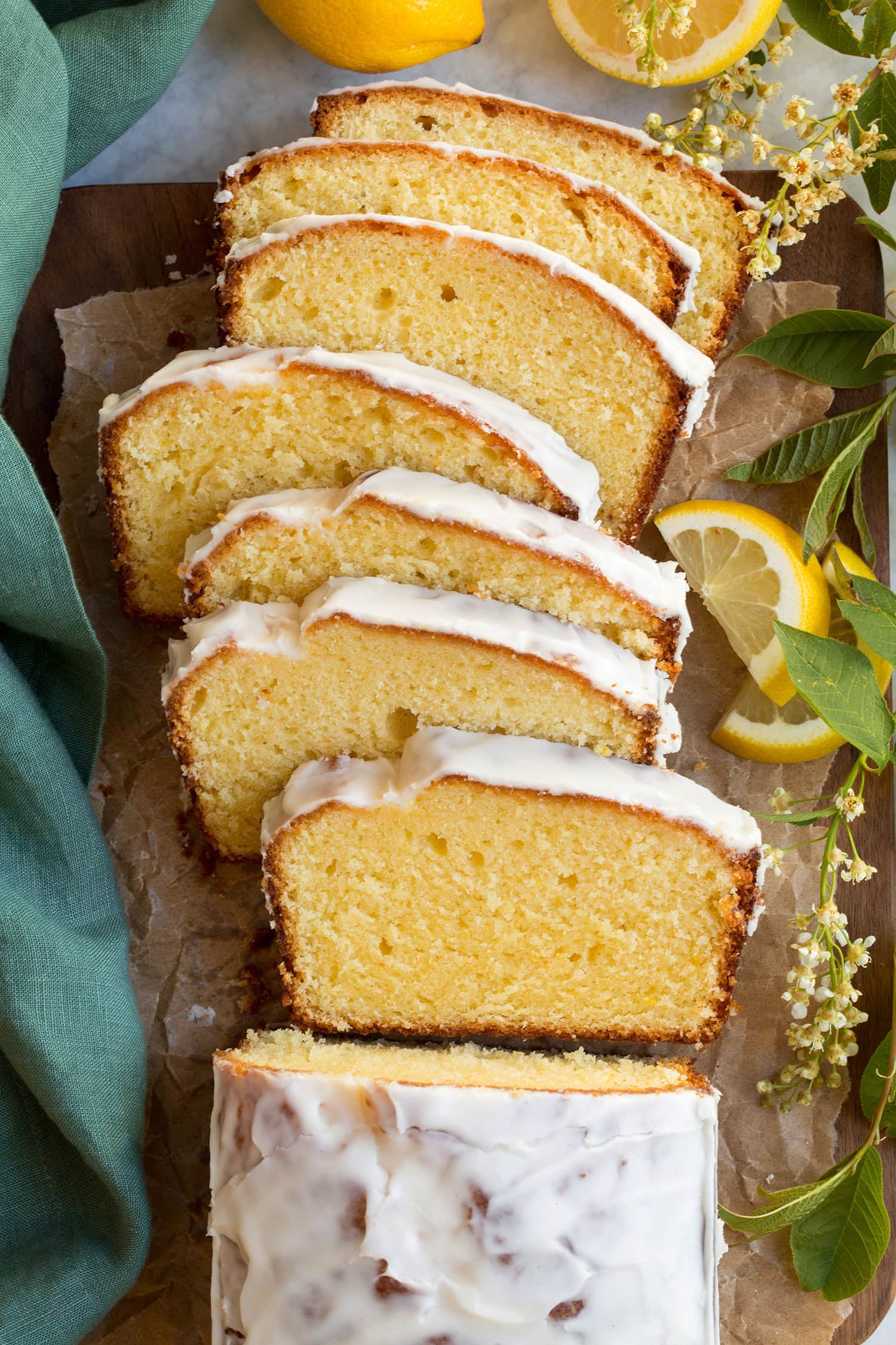 Lemon Pound Cake Lemon pound cake shown close up sliced overhead. There are lemon slices to the side and pretty green leaves and white flowers.