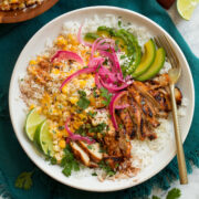 Overhead image of street corn and chicken rice bowl on a blue cloth on a marble surface.