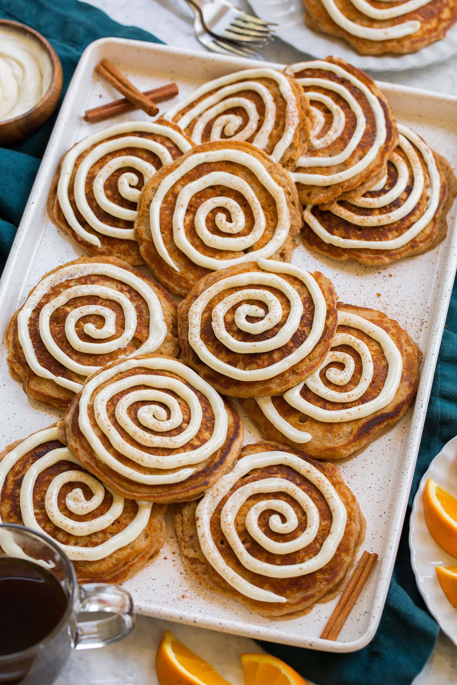 Cinnamon Roll Pancakes White baking tray full of cinnamon roll pancakes, shown over a teal cloth with oranges and coffee as serving suggestions.